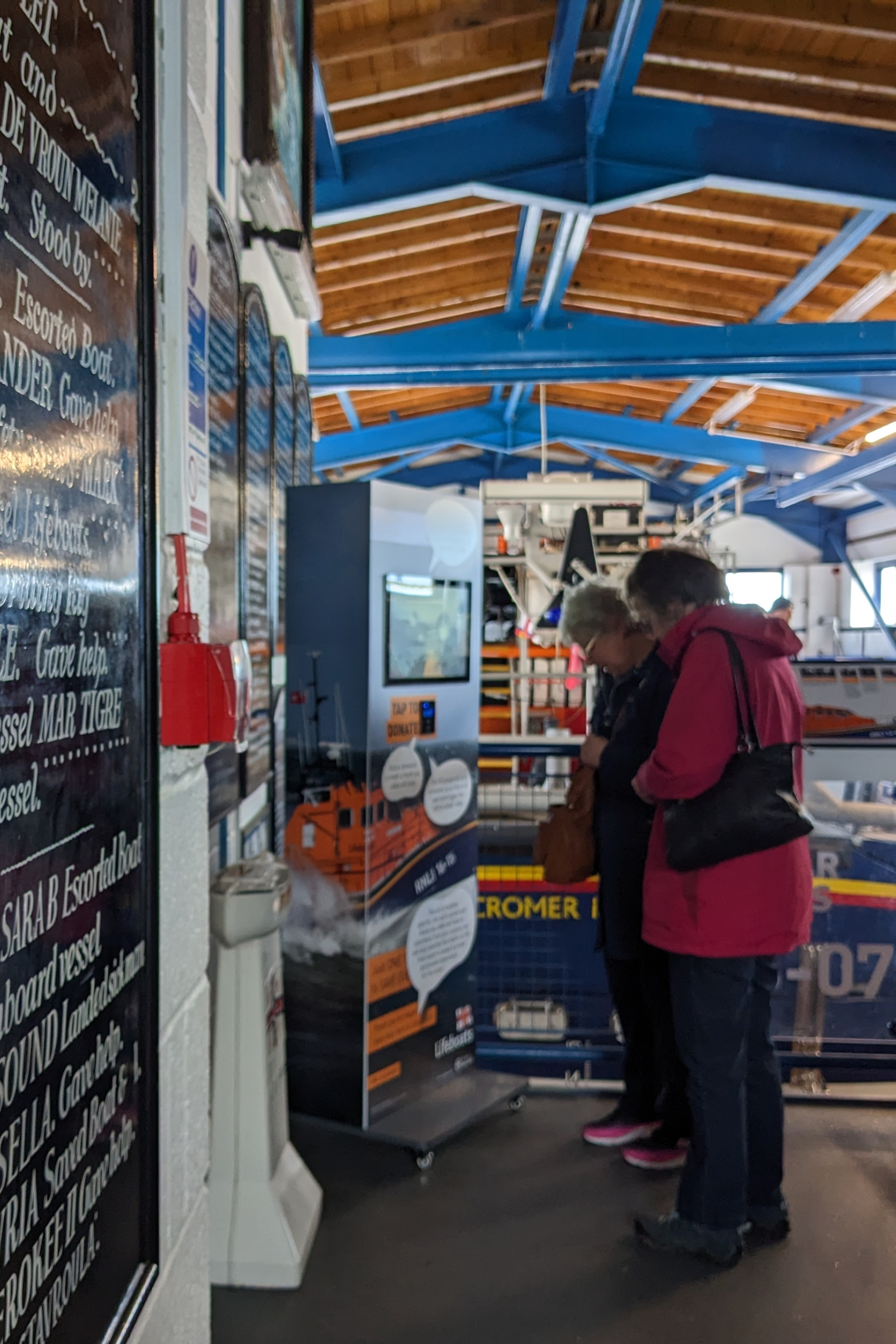 Photo of two people interacting with a Donation Triggered Video stand with RNLI branding, showing a video screen display along with a contactless payment reader.